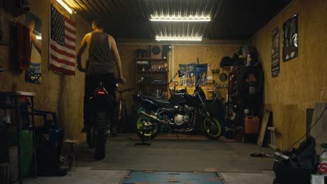 A-male-motorcyclist-with-a-short-haircut-and-a-gray-tank-top-rolls-his-motorcycle-into-his-garage-in-a-workshop-with-numerous-motorcycle-repair-tools