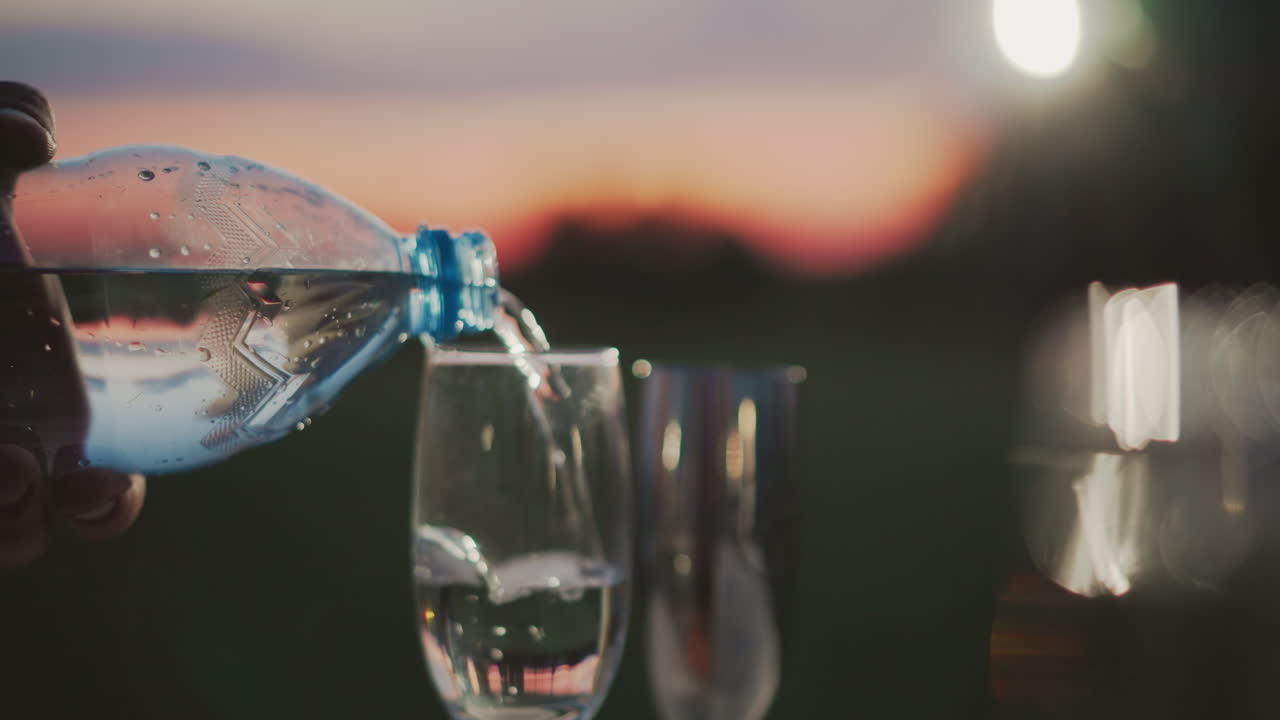 close up of water pouring from plastic bottle into glass cup against out of focus sunset bokeh lights capturing fluid motion and shimmering reflections under soft twilight sky