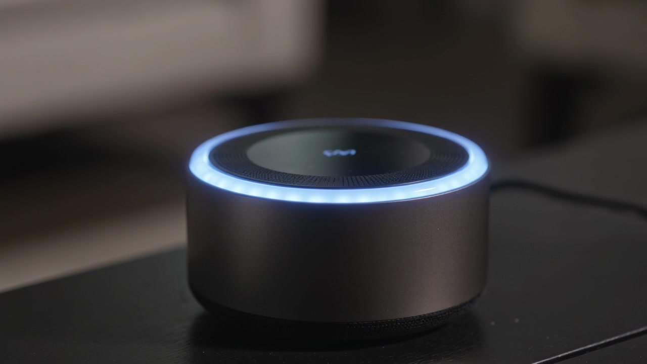 Altoparlante smart pulsante alimentato, con anello LED blu, su un tavolo di casa, che mostra il cavo di alimentazione.