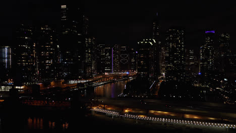 Aerial-drone-forward-moving-shot-of-traffic-movement-over-a-bridge-on-a-river-passing-through-high-rise-office-and-residential-buildings-in-Chicago,-USA-at-night-time