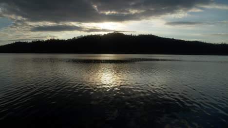 Close-aerial-flyover-of-Whiskeytown-Lake-very-close-to-the-water-that-pans-up-to-reveal-a-mountain-with-sunlit-clouds-in-the-background