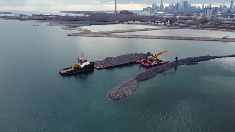 excavator on barge building breakwater from rocks with toronto skyline in background