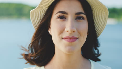 Portrait-Of-The-Beautiful-Brunette-Young-Woman-In-The-Hat-Smiling-Cheerfully-On-The-Lake-Background