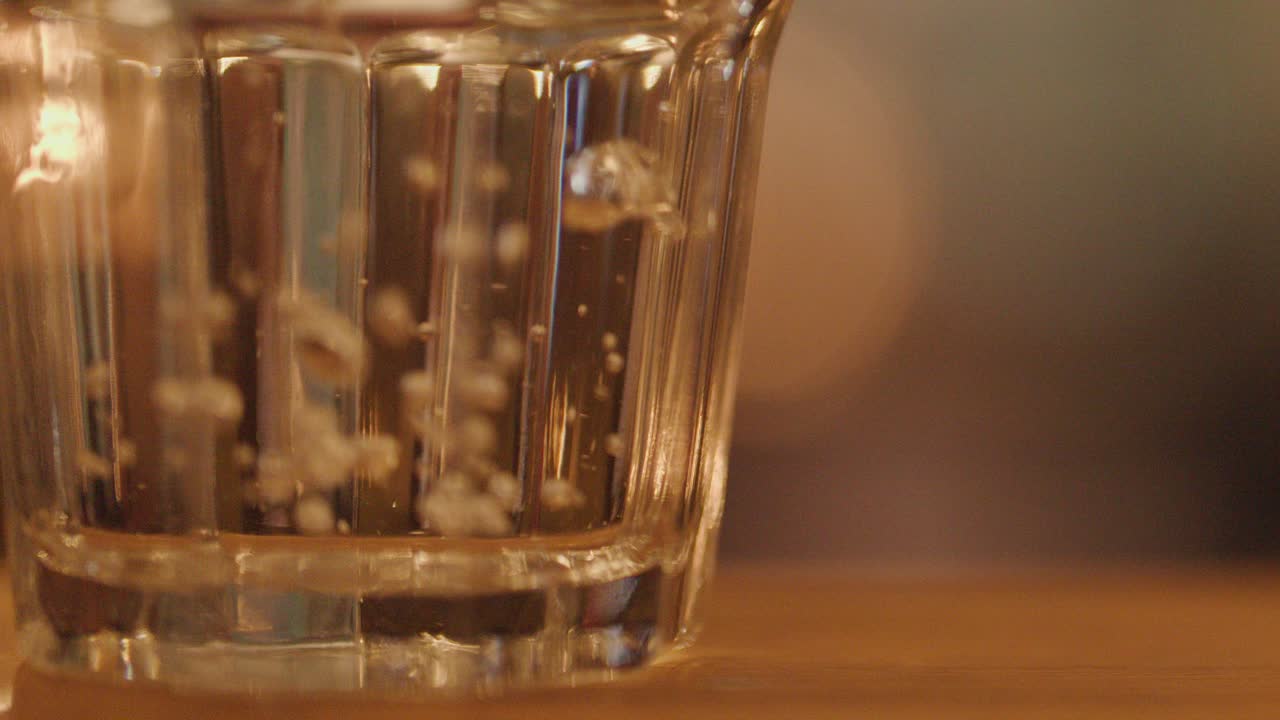 A clear glass of water on a wooden table receives an ice cube, creating splashes and bubbles. Warm, blurred restaurant lighting and shallow depth of field enhance the cozy nighttime ambiance