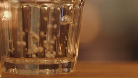 a clear glass of water on a wooden table receives an ice cube, creating splashes and bubbles. warm, blurred restaurant lighting and shallow depth of field enhance the cozy nighttime ambiance