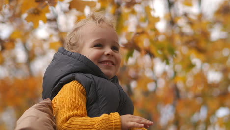 Niño-Feliz-Está-Sentado-Sobre-Los-Hombros-De-Sus-Padres-En-Un-Paseo-Por-El-Parque-En-Otoño-Retrato-De-Primer-Plano-De-Un-Pequeño-Niño-Sonriente-Feliz-Fin-De-Semana-Familiar-En-La-Naturaleza