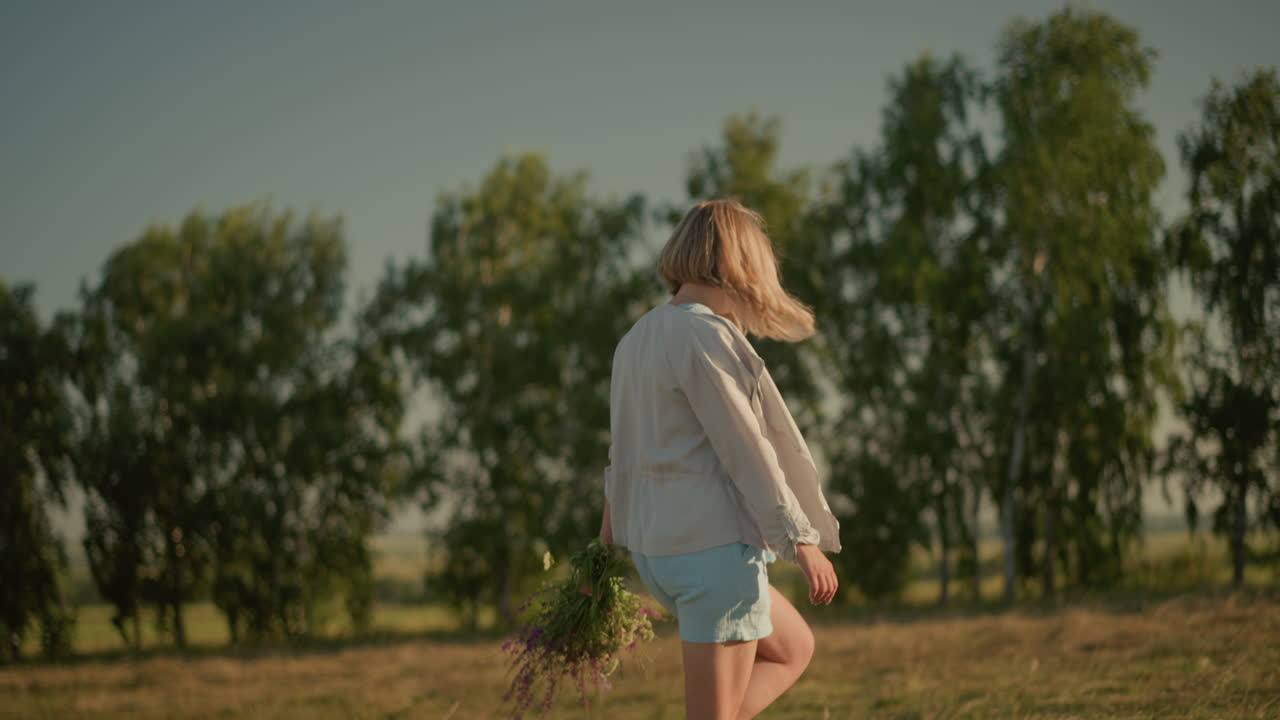 짧은 바지와 긴 팔을 가진 셔츠를 입은 여성이 은 날 손에 꽃을 들고 열린 을 걸어가고, 배경에는 흐릿한 나무와 초록색이 있습니다.