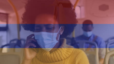 animation of flag of columbia over african american woman in face mask using smartphone in bus