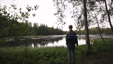 Young-man-with-camera-walks-in-forest-and-stops-in-front-of-lake,-pan