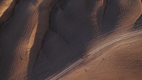 Eine-Von-Oben-Nach-Unten-Zeigende-Drohnenaufnahme-Einer-Marokkanischen-Wüste-Mit-Reitenden-Kamelen-Im-Schwachen-Abendlicht,-Das-Tiefe-Schatten-Erzeugt,-Wenn-Sie-Auf-Die-Felsigen-Wüstendünen-Treffen