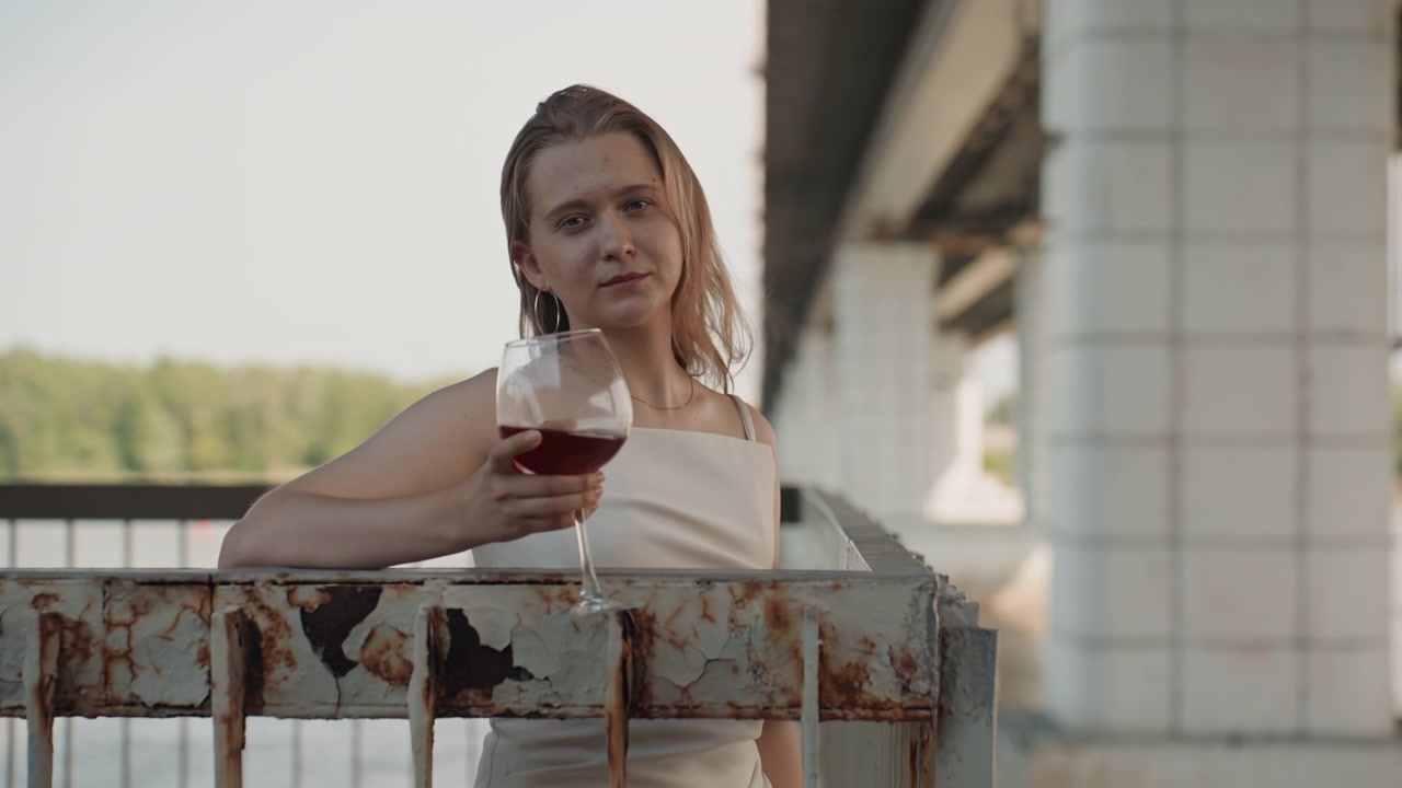 Donna in abito bianco in piedi su un vecchio ponte, che tiene un bicchiere di vino rosso e si sistema i capelli. Sullo sfondo, acqua con vegetazione al di là del fiume sotto una luce diurna soffusa.