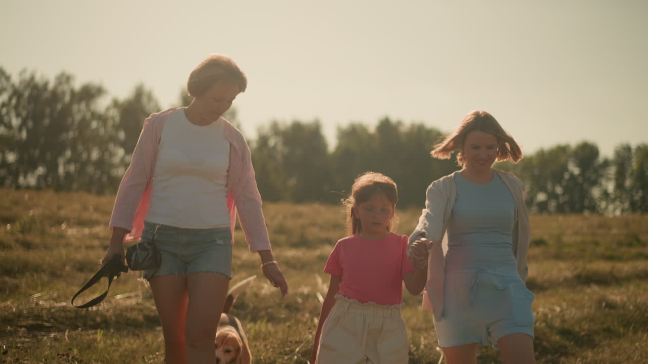 엄마의 손을 들고 있는 아이는 엄마가 따뜻하게 미소 짓는 동안 위를 쳐다보며, 이모는 줄을 들고, 먼 나무가 배경에 있는 광활한 농장에서 이 뜨는 빛의 날에 개가 앞으로 나아오도록 몸짓을 한다.