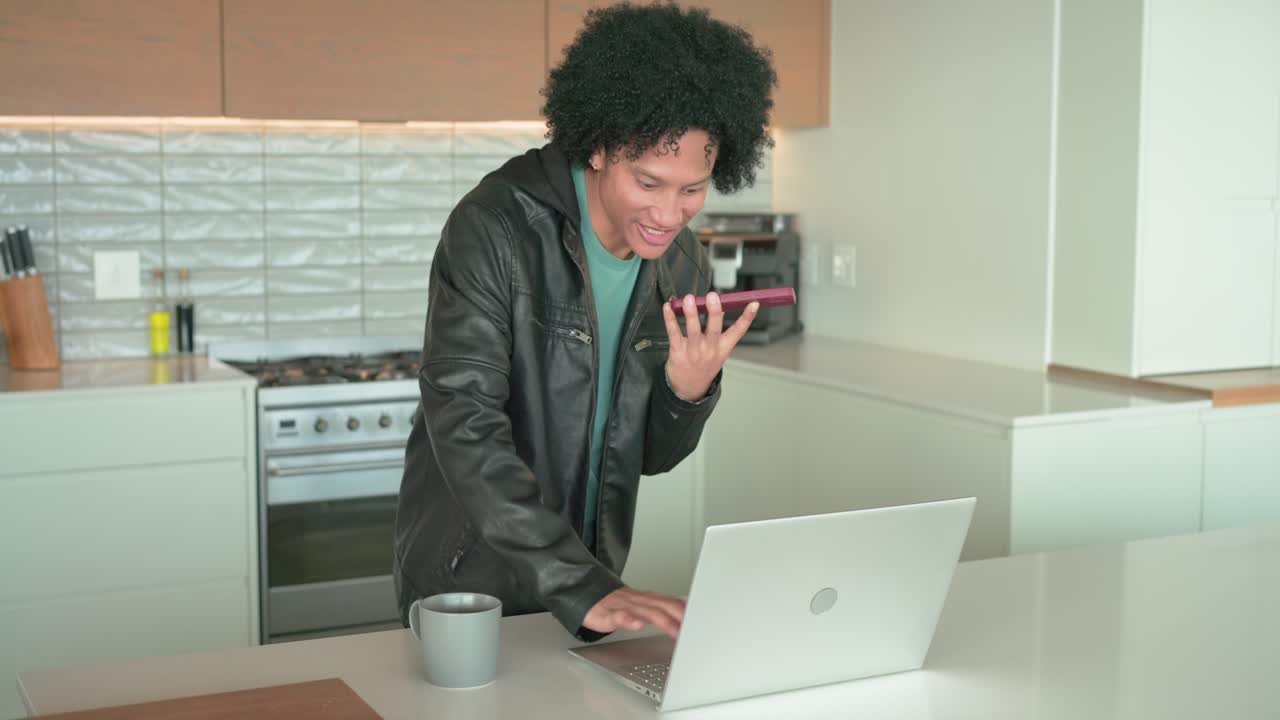 Incoming call motivating African American man speaking on phone, typing on laptop at kitchen island