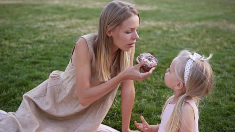 Portrait-Of-Mother-And-A-Small-Daughter,-Spends-Time-Together-In-A-City-Park-On-A-Picnic-They-Bite-A-Tasty-Donut-From-Sides-Together