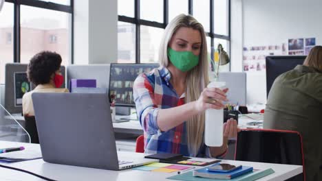 mujer de negocios caucásica con máscara facial desinfectando las manos usando una computadora portátil en una oficina moderna