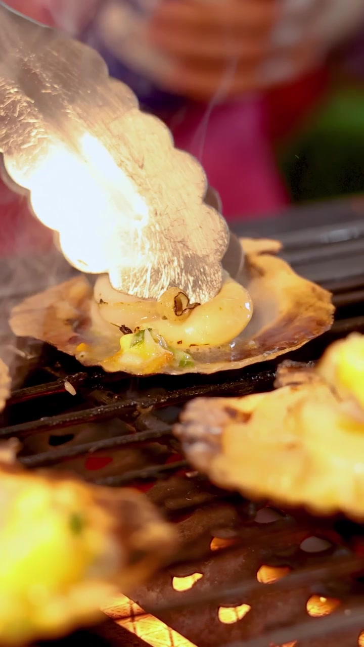 Tongs turning scallops on shells over open grill, vibrant lighting, shallow depth, street food scene