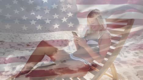 bandera estadounidense ondeando contra mujer caucásica usando un teléfono inteligente sentada en una silla de cubierta en la playa