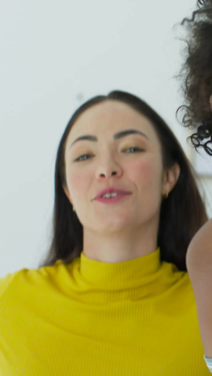 Vertical video: Smiling curly woman turning, friend yellow top coming from left to greet by window