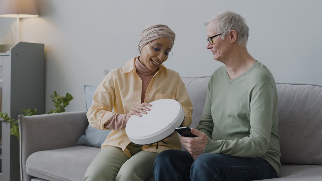 집에서 함께 소파에 앉아 로봇 진공 청소기를 사용하는 방법을 노인에게 설명하는 중년 아랍 여성
