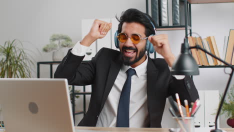 indian businessman wearing headphones listening music dancing after working on laptop at home office