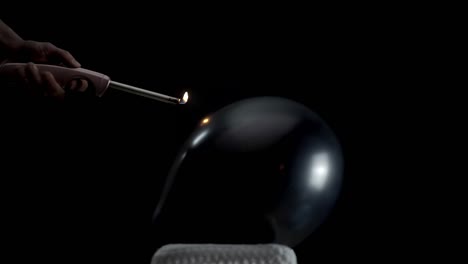 a black balloon lays still against a black background. a hand holding a lighter moves closer and closer, building tension until the balloon pops in a dramatic, slow-motion moment of suspense.