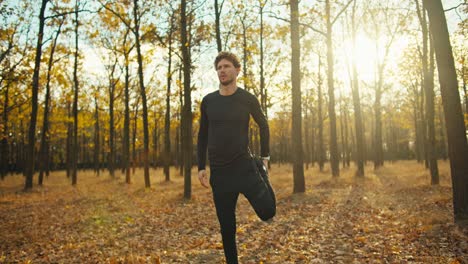 Un-Hombre-Seguro-Y-Serio-Con-Cabello-Rizado-Con-Un-Uniforme-Deportivo-Negro-Está-Calentando-Y-Luego-Mira-Su-Reloj-Y-Comienza-A-Trotar-En-El-Bosque-Otoñal-De-La-Mañana-En-Otoño.