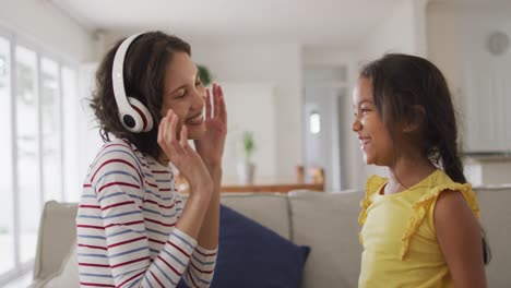 Happy-hispanic-mother-wearing-headphones-listening-to-music-and-daughter-sitting-on-sofa