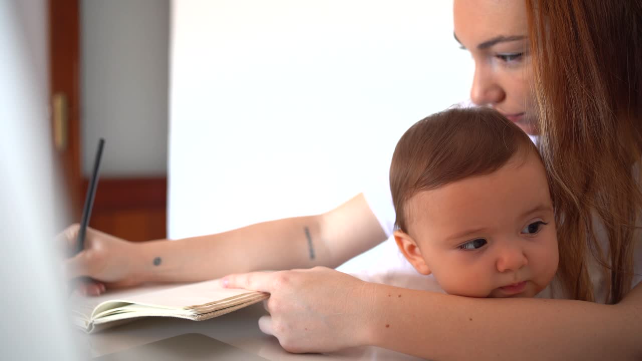 Young mother hugging baby on laps and writing in copybook