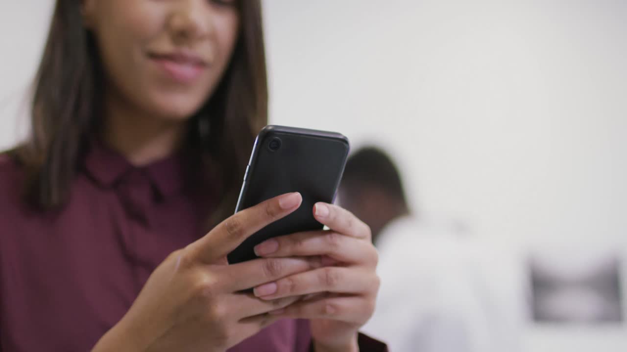 mujer de negocios biracial sonriente usando un teléfono inteligente con colegas en el fondo en una oficina moderna