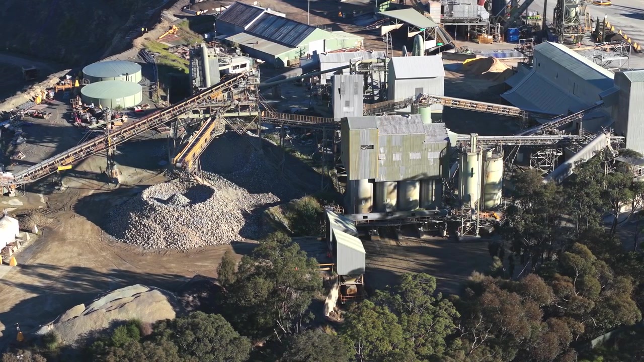 A dynamic aerial shot captures Montrose Quarry in Melbourne, revealing rugged machinery, conveyor belts, and raw aggregate piles under crisp daylight, trees and industrial structures. Aerial footage.
