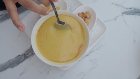 a hand stirring a bowl of yellow soup with a spoon