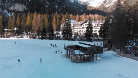 Panorámica-Aérea-Lenta-De-Gente-Caminando-Cerca-De-Una-Antigua-Estructura-De-Madera-Sobre-Un-Lago-Congelado-Braies-En-Italia