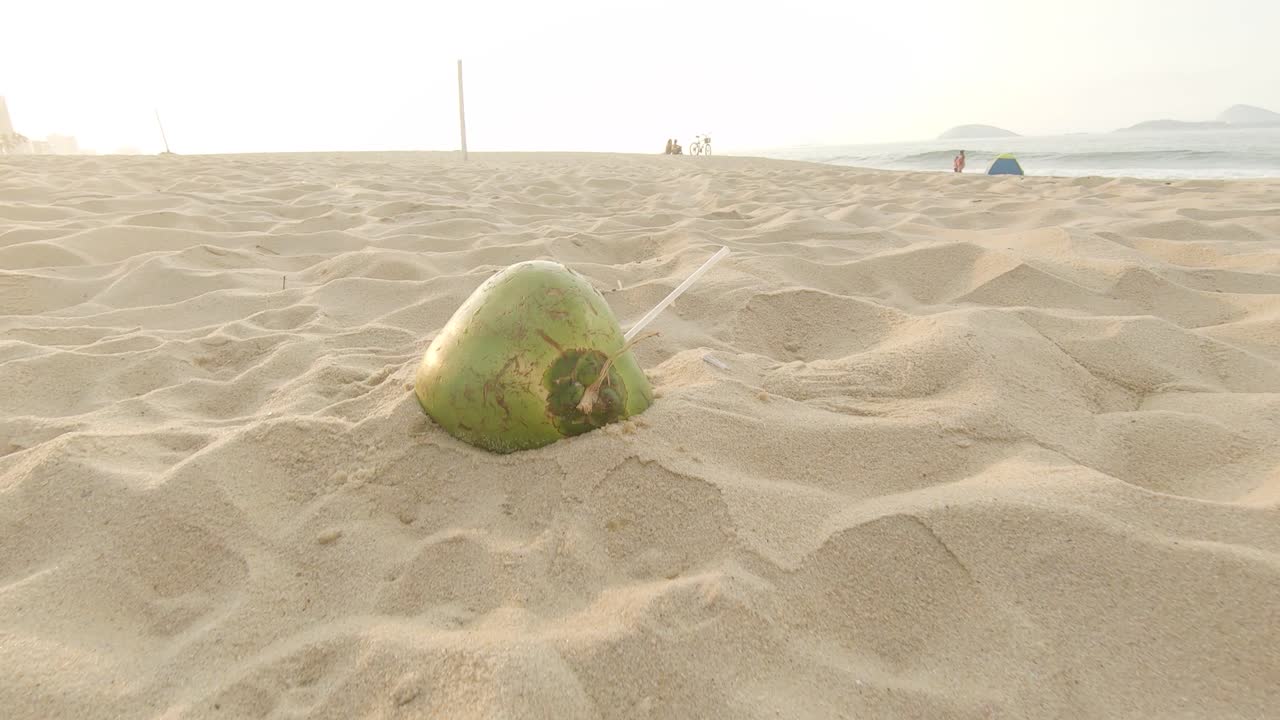해안가에 앉아 백그라운드에서 지나가는 사람들과 함께 일출 해변에 플라스틱 빨대가 튀어나온 빈 코코넛 껍질 뒤에 남겨졌습니다.
