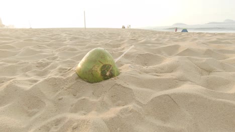 Left-behind-empty-coconut-shell-with-a-plastic-straw-sticking-out-on-a-beach-at-sunrise-with-people-sitting-at-the-waterfront-and-passing-by-in-the-background