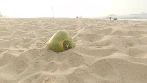 dejado atrás una cáscara de coco vacía con una pajita de plástico que sobresale en una playa al amanecer con gente sentada en el paseo marítimo y pasando por el fondo
