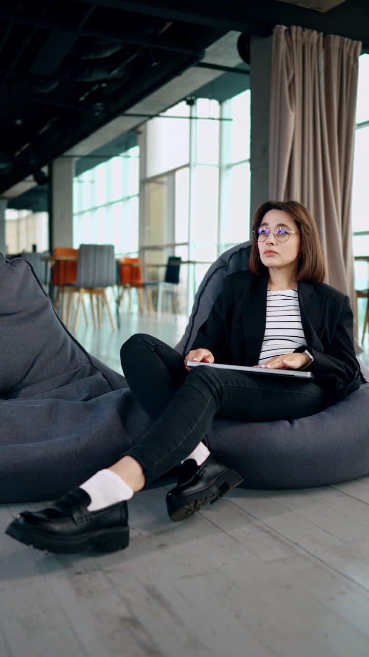 Young woman sits in the bean bag chair in a spacious office. Lady is holding a laptop on her laps. Remote work concept. Low angle view. Vertical video.