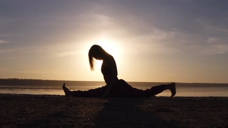 young woman doing split, yoga posture outdoors on the beach. beautiful silhouette of athlete. sunset