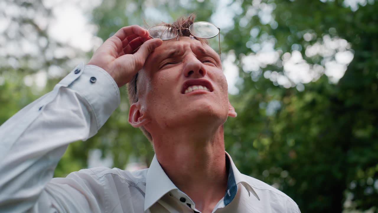 Close view of young man in white shirt adjusting glasses while observing trees in outdoor park setting, surrounded by lush greenery with natural daylight creating thoughtful expression