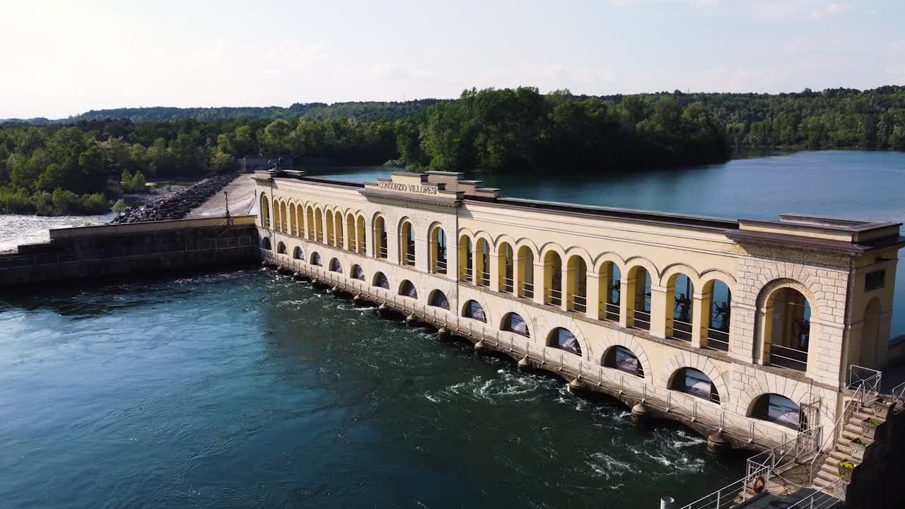 Ripresa aerea con drone di una storica centrale idroelettrica dei primi del '900 con facciata gialla ad arco che attraversa un fiume turbolento, circondata da una foresta verde in Lombardia, Italia settentrionale.