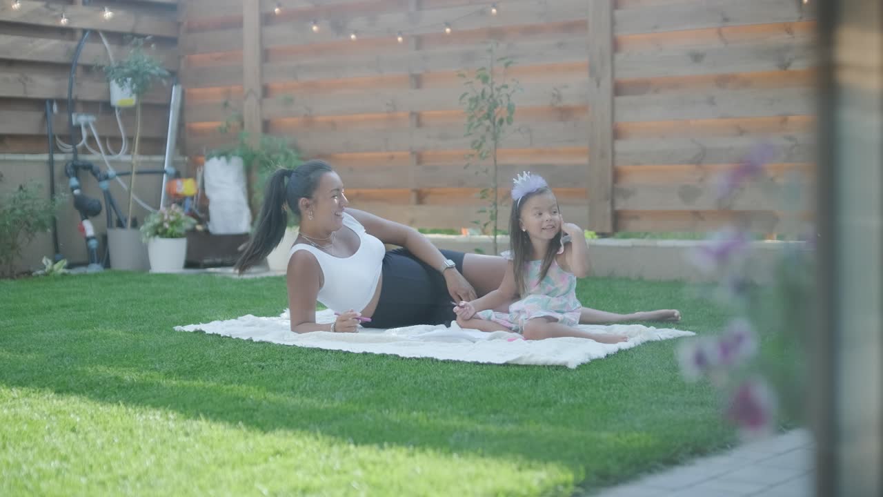 A mother and her young daughter sit on a blanket in the backyard, engaging in a drawing activity together. The outdoor scene captures a peaceful family moment, fostering creativity and bonding.
