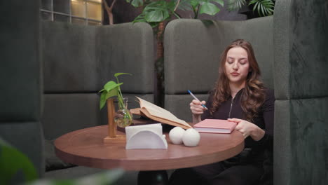 lady in black outfit adjusts sleeve while seated at round table with closed pink book and open brown book, preparing to read or write in cozy green booth with relaxed focused expression