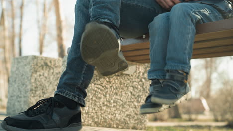 close-up of an adult and a child sitting closely together on a bench, with the child playfully dangling his legs and the adult moving his crossed legs, with a blurred background of trees