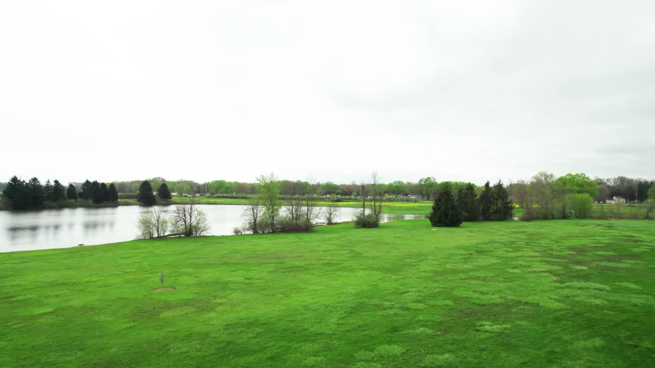 снимок с воздушного дрона, пролетающего над полем для диск-гольфа в направлении озера и шоссе, которое находится вдалеке, ранней весной с яркими зелеными цветами