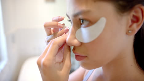 Focused-biracial-teenage-girl-putting-on-under-eye-masks-in-bathroom,-copy-space,-slow-motion