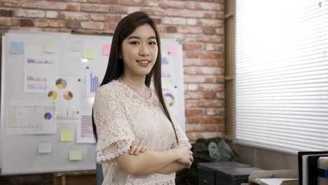 pretty confident asian female manager is smiling at the camera with crossed arms in a bright loft office at daytime.