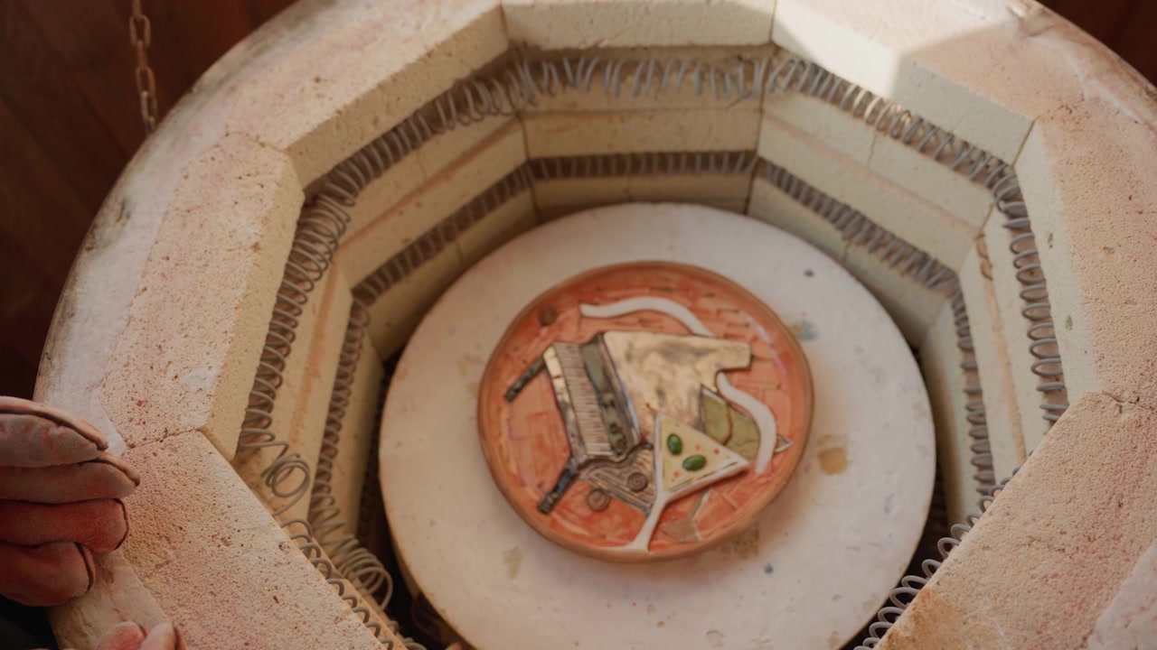 Primo piano dell'interno di un forno con piatto in ceramica, vasaio che ispeziona un piatto di terracotta smaltato sullo scaffale del forno, mani sul bordo, in raffreddamento dopo la cottura ad alta temperatura, con isolamento in mattoni e catena appesa visibili.