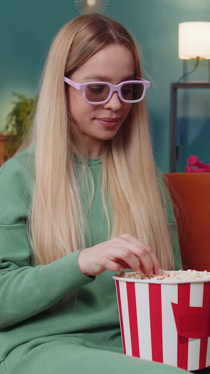 Giovane donna seduta sul divano che mangia popcorn e guarda un'interessante serie TV o una partita sportiva online a casa