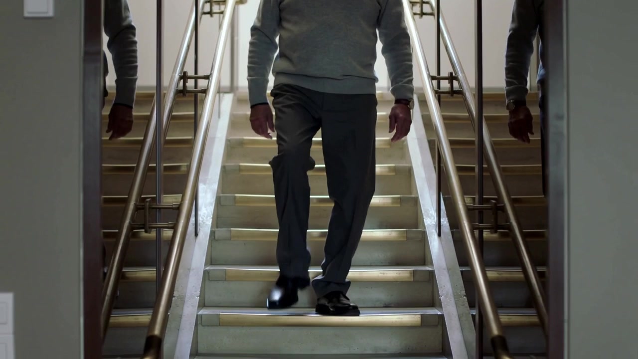 Voksen mand i sweater sætter højre fod og går ned ad en lige trappe, hvor spejle og gelændere er synlige.