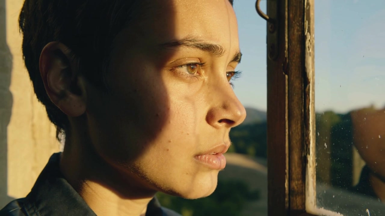 Standing woman gazing out wood-frame window at home, sun catching tear and reflection, shirt