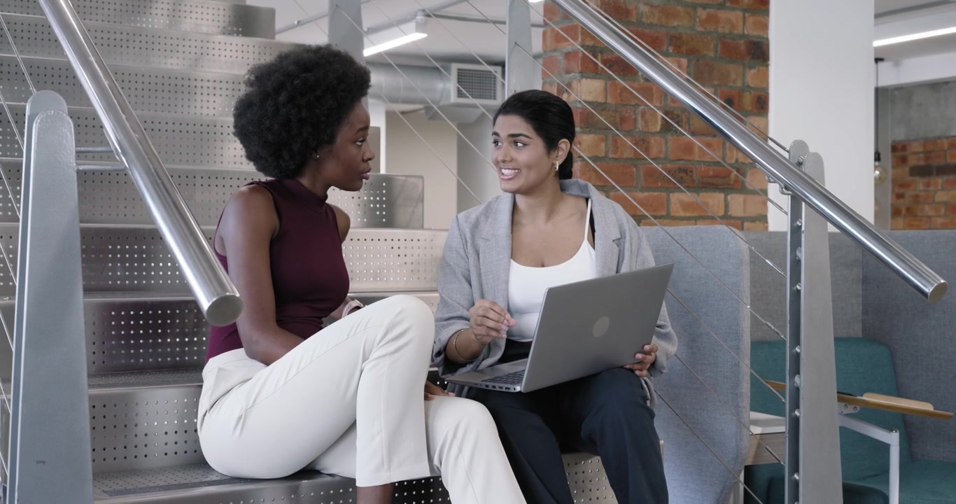 Diverse women coworkers working on lap laptop on metal stairs, right typing left leaning gesturing